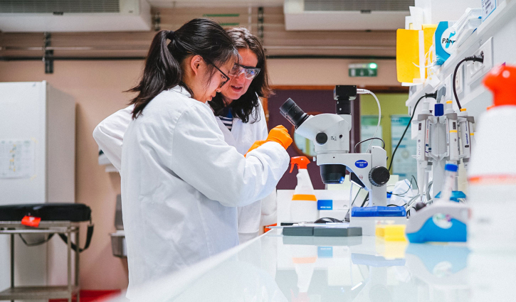 Two women in white lab coats stand at a lab bench with a microscope, examining something the camera can’t see