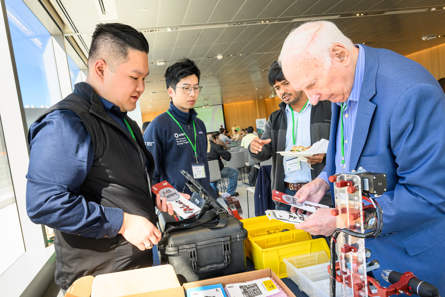 Russel Bradley and Ray Stata holding pamphlets confer over a table bearing a small robot, among other things