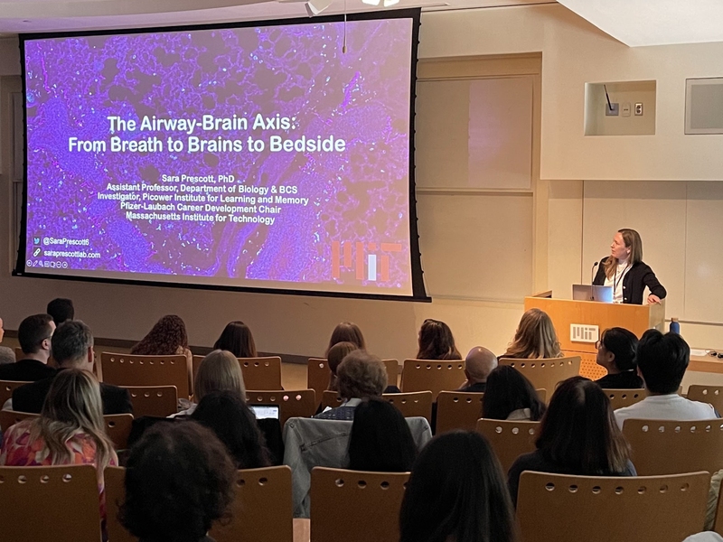 Sara Prescott stands at a podium as she begins delivering her talk to a seated audience. The title of her talk is on a slide on the big screen. 