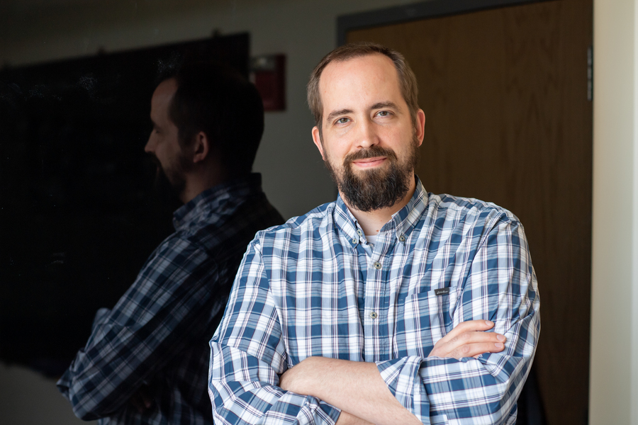 Matt Shoulders, wearing a blue-and-white plaid shirt, stands with his arms crossed
