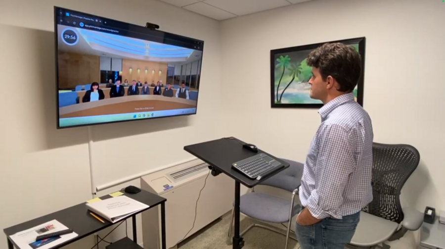 A graduate student stands in the Communication Studio, pitching to a virtual audience on a mounted screen.