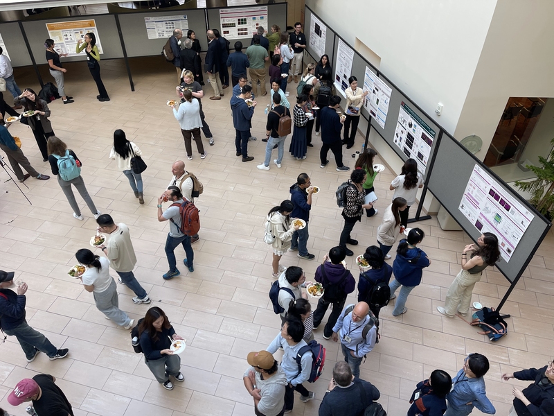 Overhead view of the atrium of MIT's Building 46 shows a crowd, many with plates of food, milling about. Research posters line boards along the walls of the atrium.