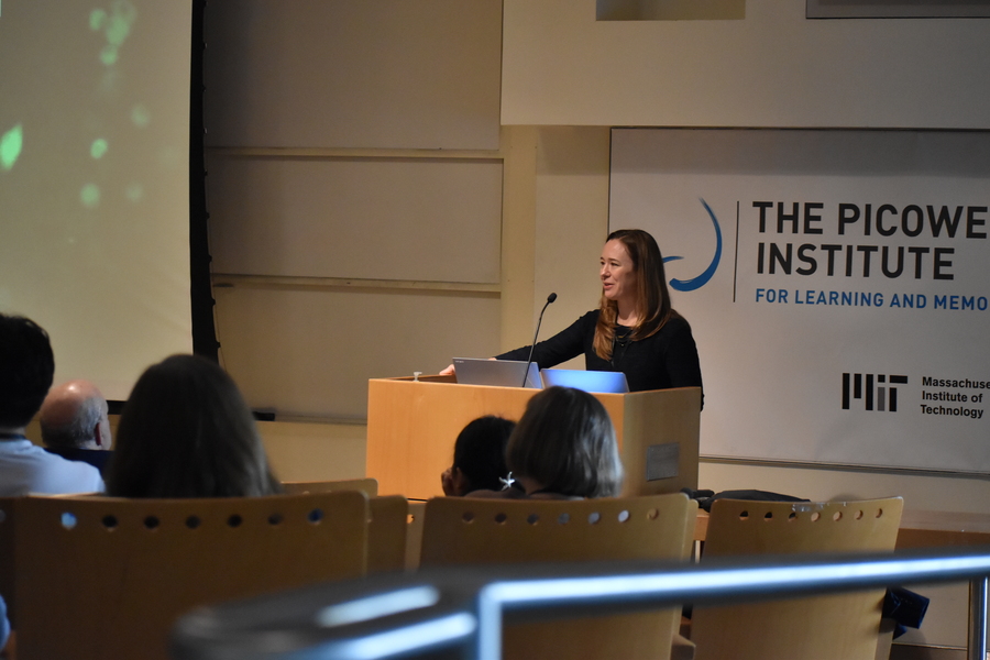 Sara Prescott speaks from behind a lectern with audience members in the foreground and a Picower Institute banner in the background
