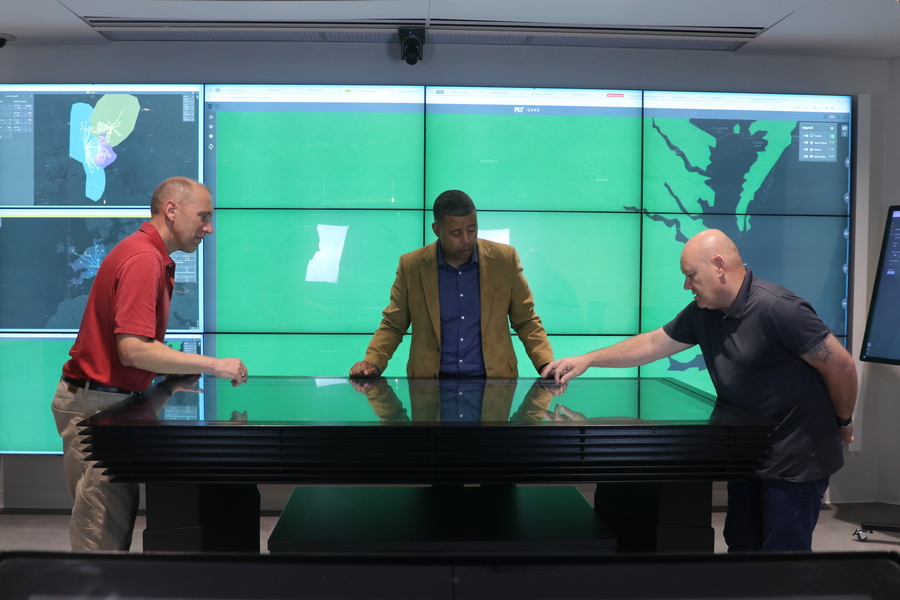 Three men stand over a glass-topped table in front of a bank of nine monitors.