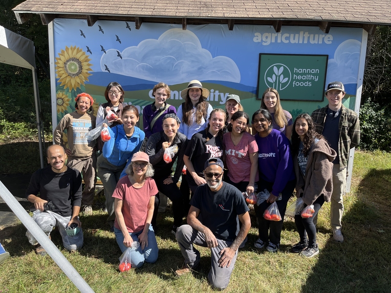 16 people, several holding up clear plastic bags containing tomatoes, pose for a photo at a farm.