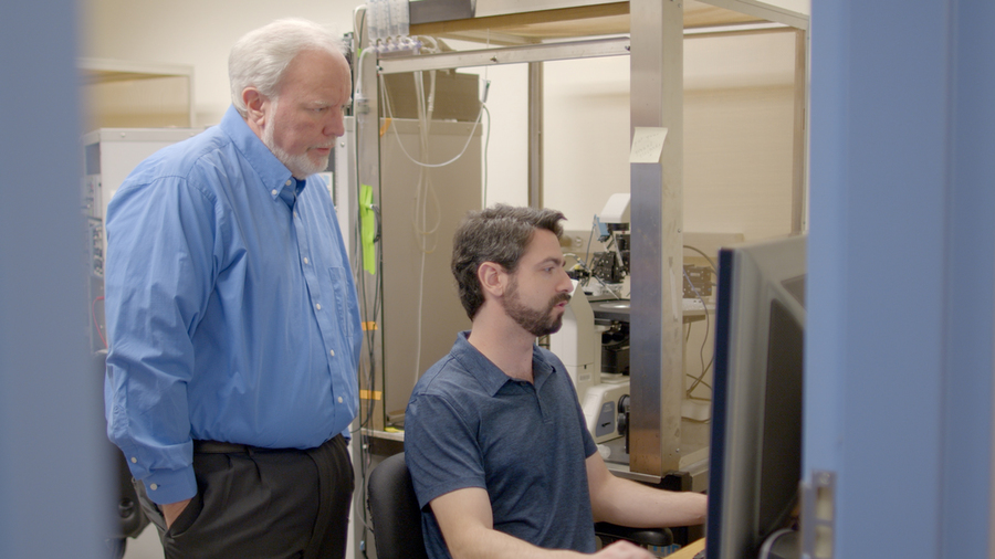 Andres Crane sits at a computer workstation in a lab. Troy Littleton stands behind him, looking at the screen