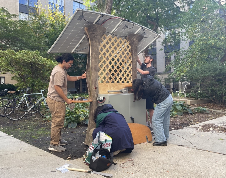 Four students installing solar-powered charging station comprising a metal electronics cabinet, two tree trunk-like pillars, wooden lattice, and four solar panels