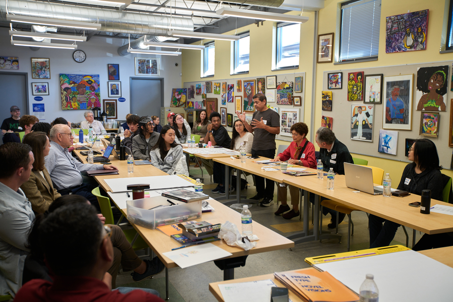 A conference-style room filled with people seated at tables. One of the people is standing and talking. The walls are covered in art.
