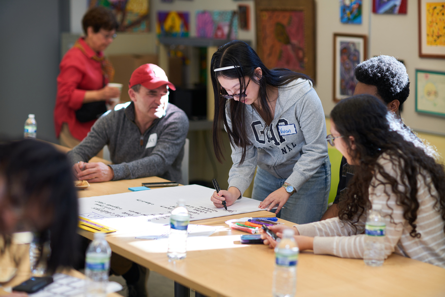 Four people are seated around a large piece of paper. One of them is writing on the paper.