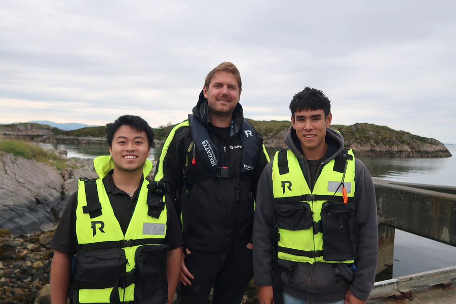 Tony Tang, Sveinung Ohrem, and Beckett Devoe pose wearing life jackets on a dock with fjords in the background.