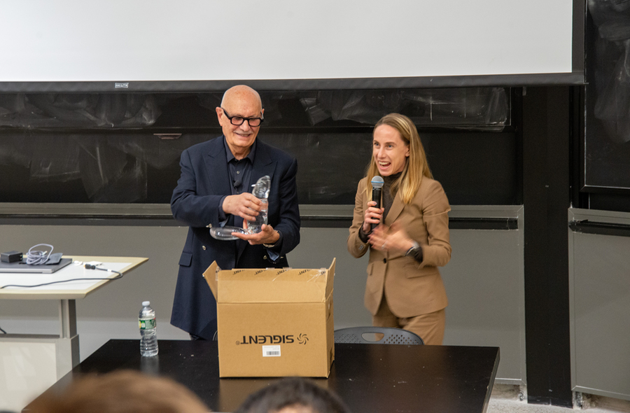 Diran Apelian and Polina Anikeeva stand behind a table in front of a blackboard. Apelian holds a glass beaver sculpture and Anikeeva holds a microphone