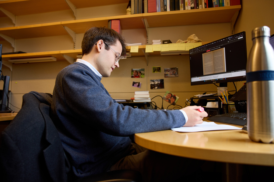 Alex Busch sits at desk facing window with pen in hand while reading and writing.