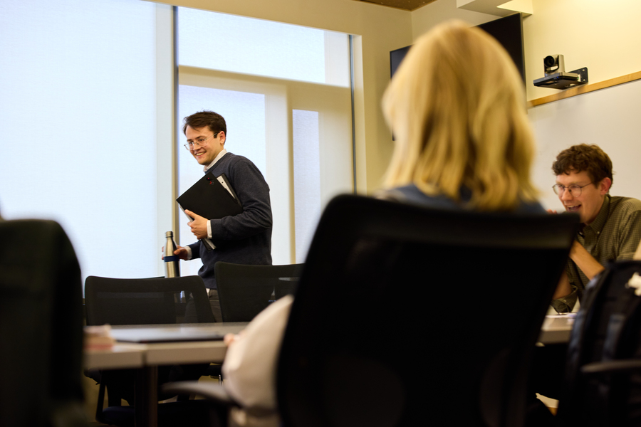 Alex Busch walks to chair while holding notebook and water bottle. Professor is seated to his left.