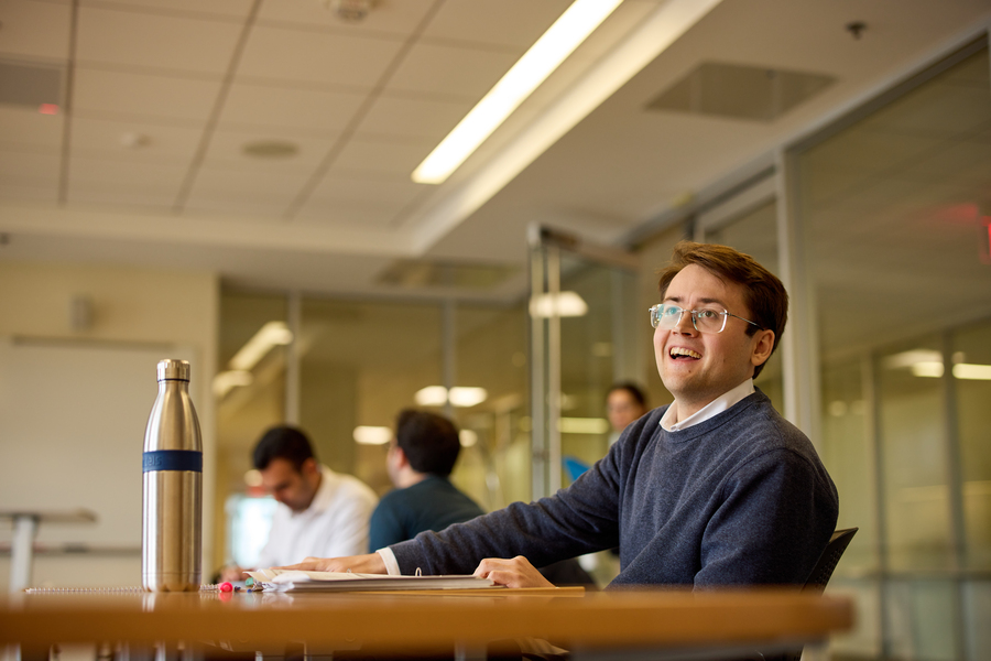 In foreground, Alex Busch sits behind a table. Three out-of-focus students are visible in the background