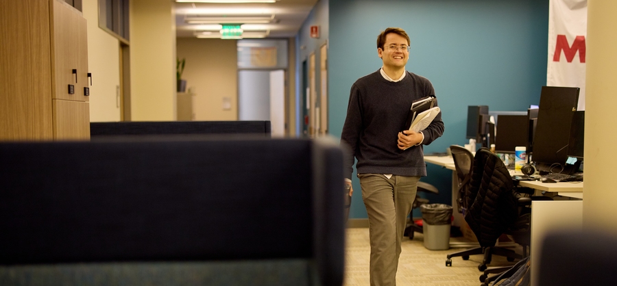 Alex Busch carries books and papers walking down a hallway by cubicles and desks