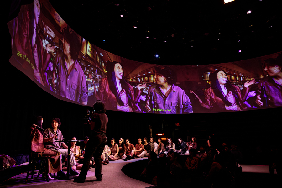 Two actors sit at a small round table, their images projected multiple times on a large circular screen above them. Some 20 others sit on the floor in a semicircle behind them.