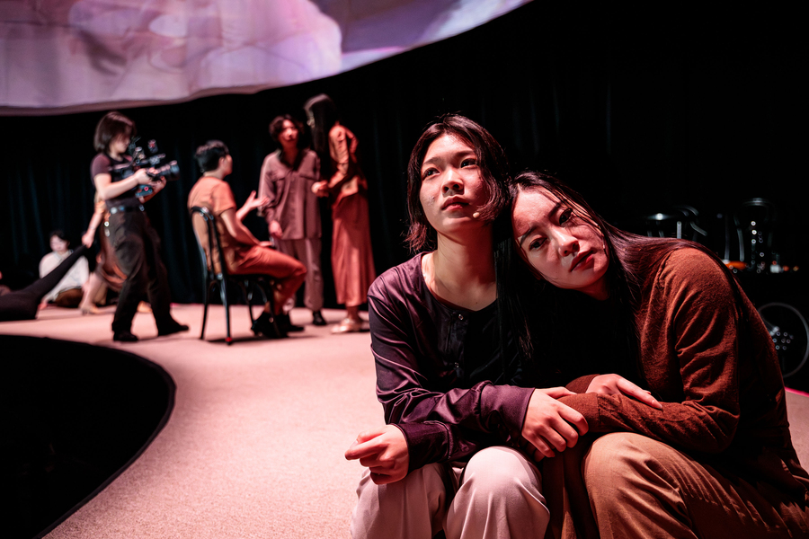 Two actors sit on stage in the foreground. Three more actors and a camera operator are out of focus in the background.