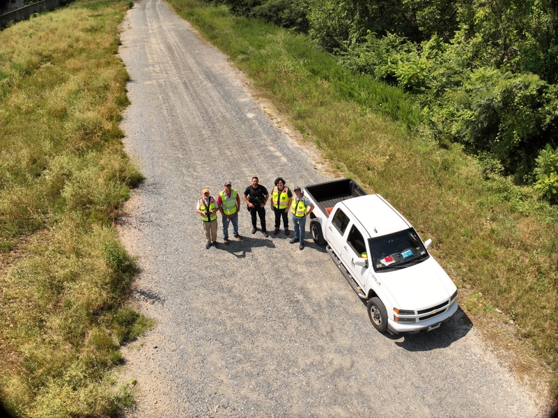 Aerial image of Diego Tempkin standing next to a pickup truck on a gravel road by a grassy area with 4 co-workers in transit authority vests.