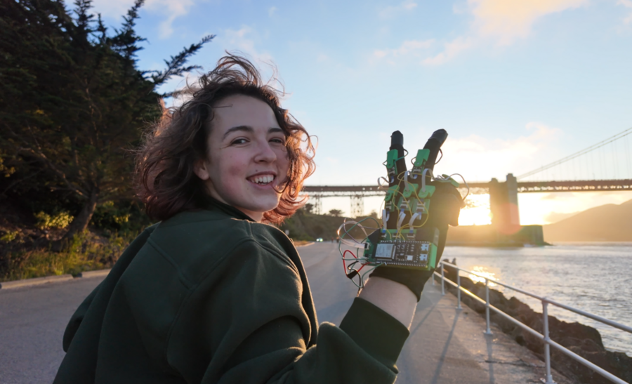 Freesia Gaul stands next to water and a large bridge, looking over her shoulder at the camera, holding up her hand wearing a haptic glove