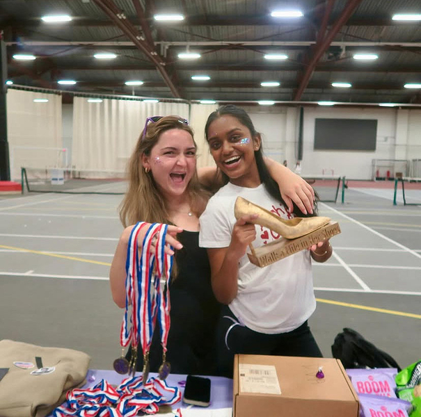 Two students in the gym. One is holding ribbons, the other a wooden high heel shoe, and both have some face paint