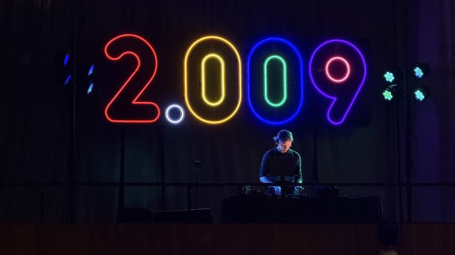 On a dark stage, a DJ stands in front of illuminated neon lights in the shape of the numbers 2.009