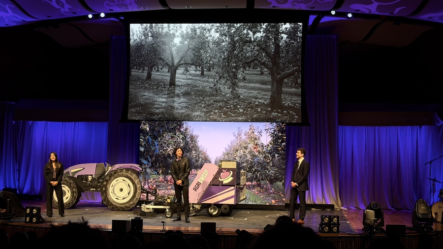 Three students on a stage present a device attached to a tractor, with an apple orchard projected behind them.