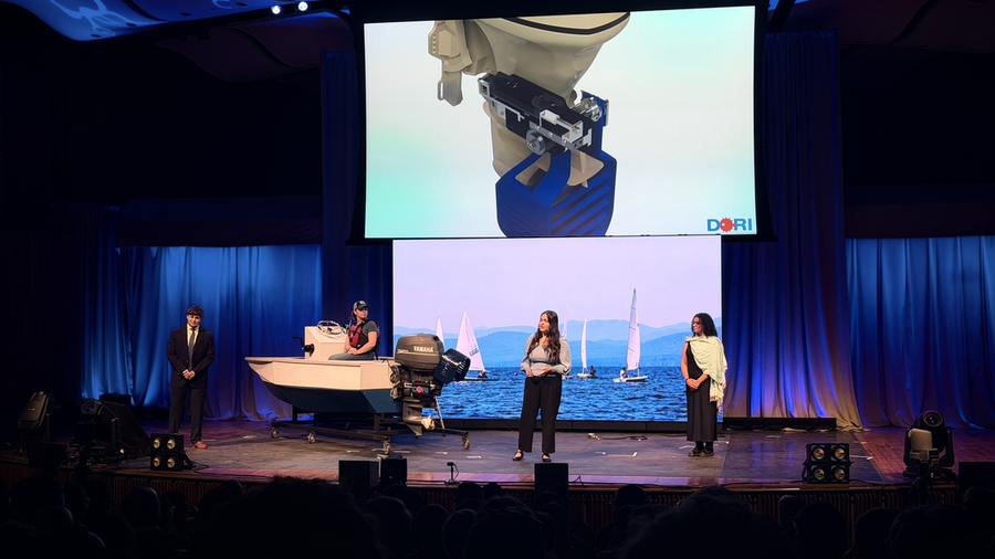 Three students stand on a stage, while a fourth sits in a motorboat on a trailer. Their device is projected on the screen behind them.