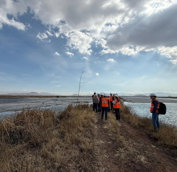 A dozen or so people in orange safety vests and hardhats gather on a point of land surrounded by water
