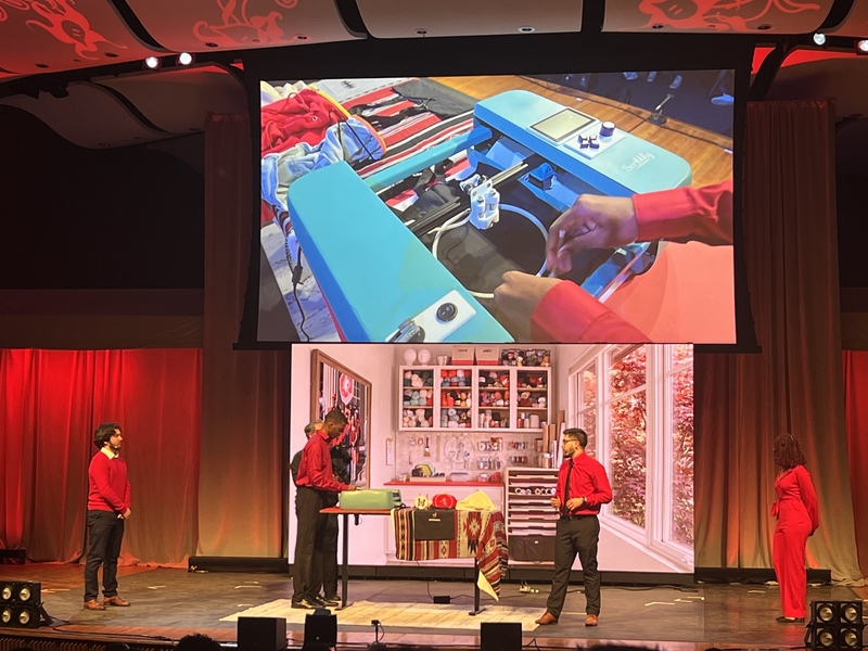 Students on a stage demonstrate a machine, with a close-up projected overhead