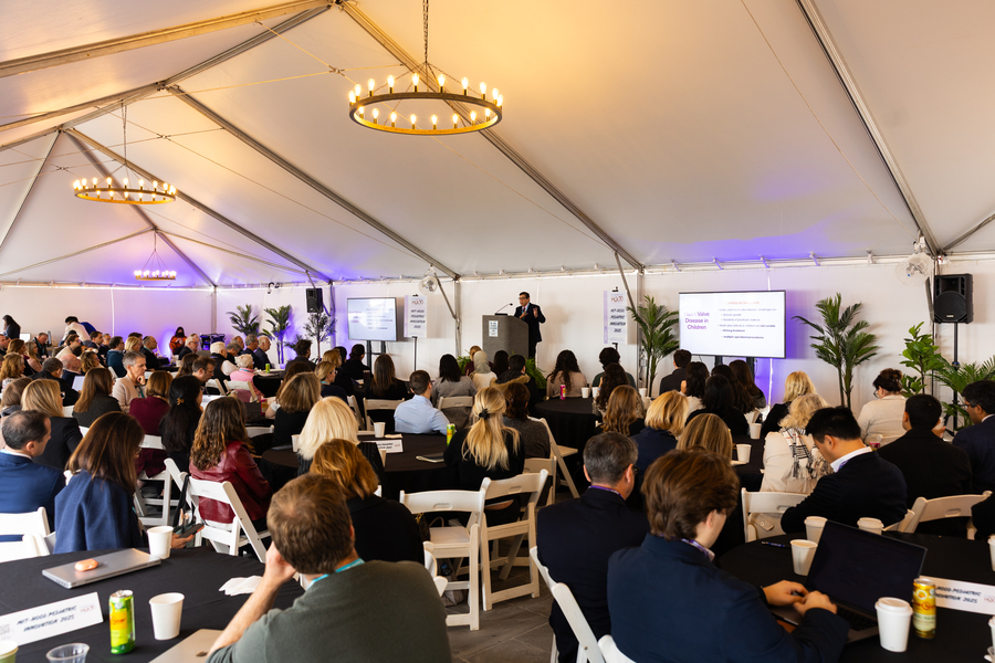Joe Frassica addresses scores of people sitting at round tables under a canvas structure