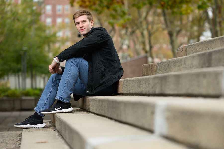 Taylor Hampson, wearing jeans and a light jacket, sitting outdoors on concrete steps with trees and a brick building in the background
