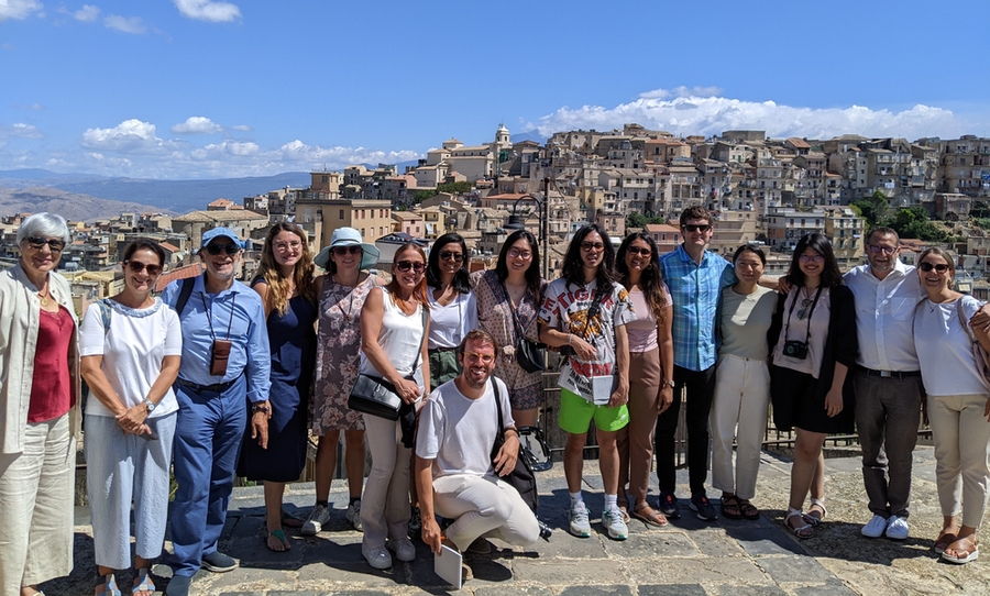16 people pose on a plaza overlooking an Italian village with mountains in background
