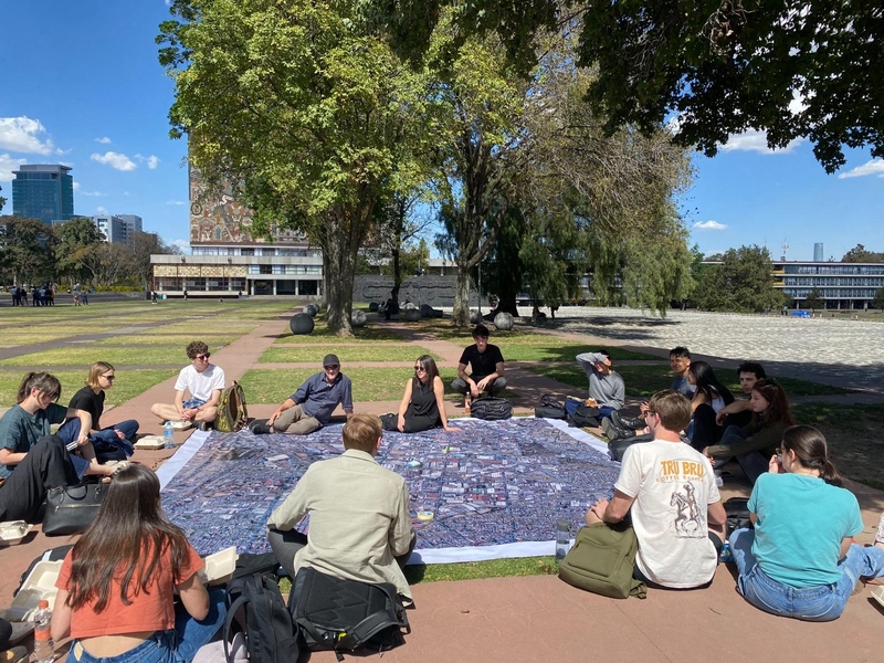 16 people sit around a large blanket in a grassy area on the Universidad Nacional Autónoma de México campus
