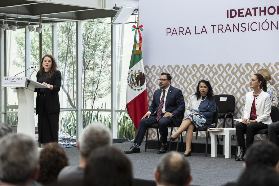 Sarah Williams speaks at a lectern indoors while three people seated on stage look on