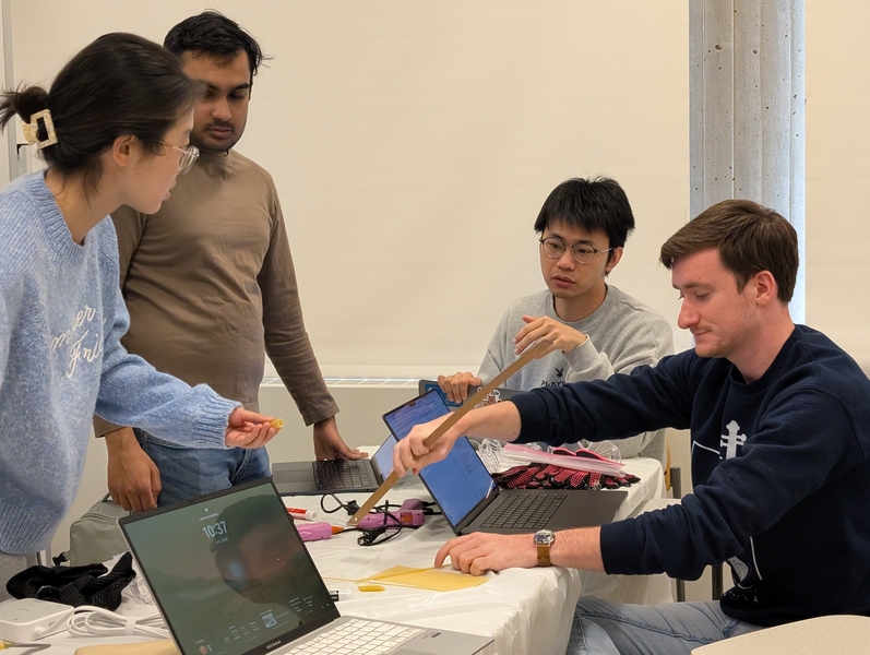 Four students gather around a table, collaborating on a hands-on project with laptops and materials.