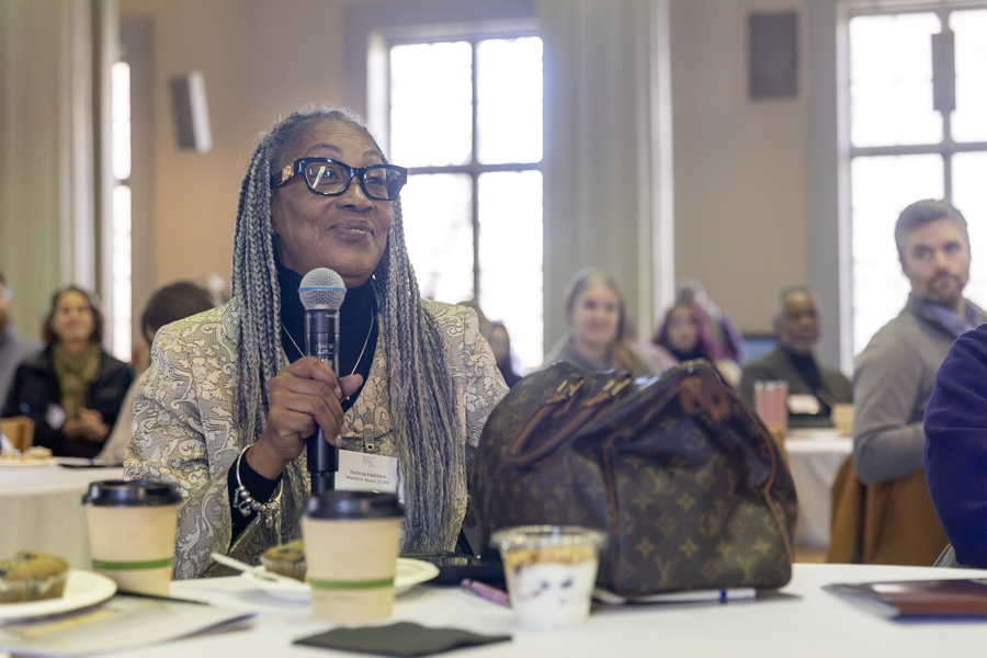 A woman with long dreadlocks seated at a table with others at tables behind her speaks into a microphone