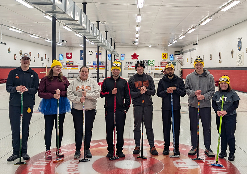 8 members of MIT's Curling team pose standing in a hockey rink.
