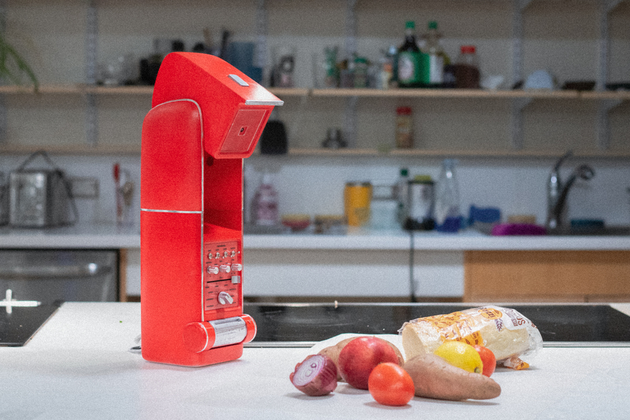 On a counter, a red Kitchen Cosmo device points its camera at assorted vegetables