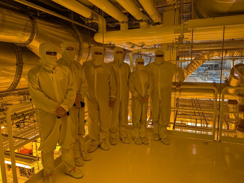 Six people in white bunny suits stand on a cleanroom fan deck under golden light.