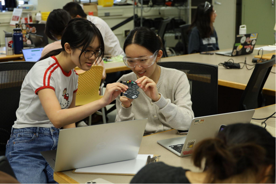 Two students seated at a table manipulate an oddly shaped circuit board