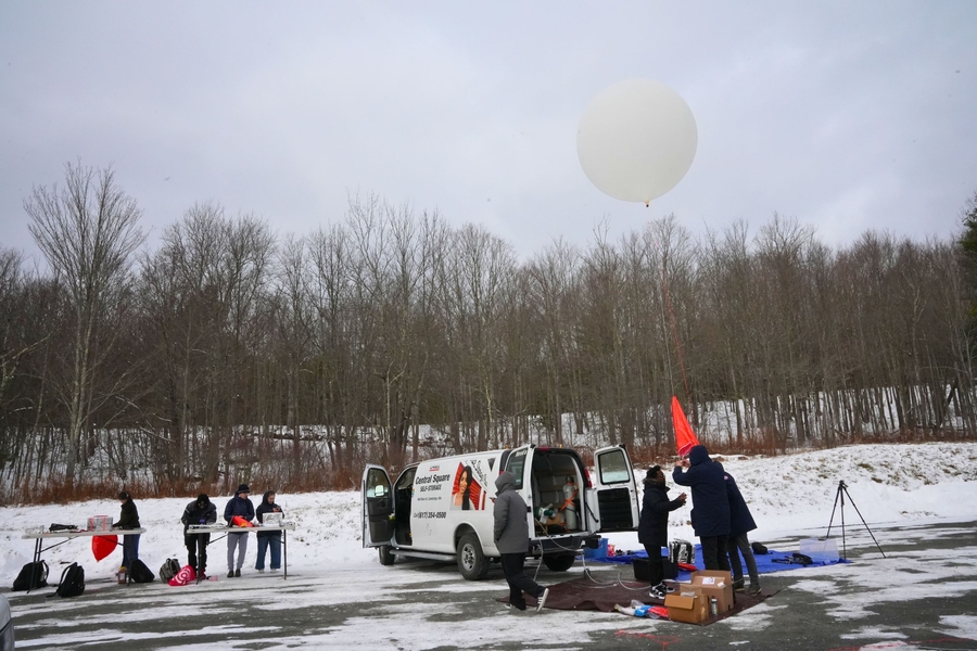People launching a large white weather balloon in a winter landscape with trees and a parked van.