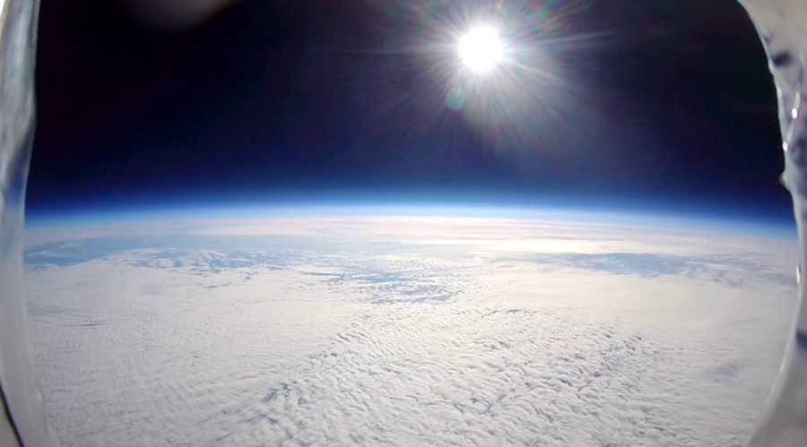View of Earth from the stratosphere, with clouds below and the sun shining in a dark blue sky.