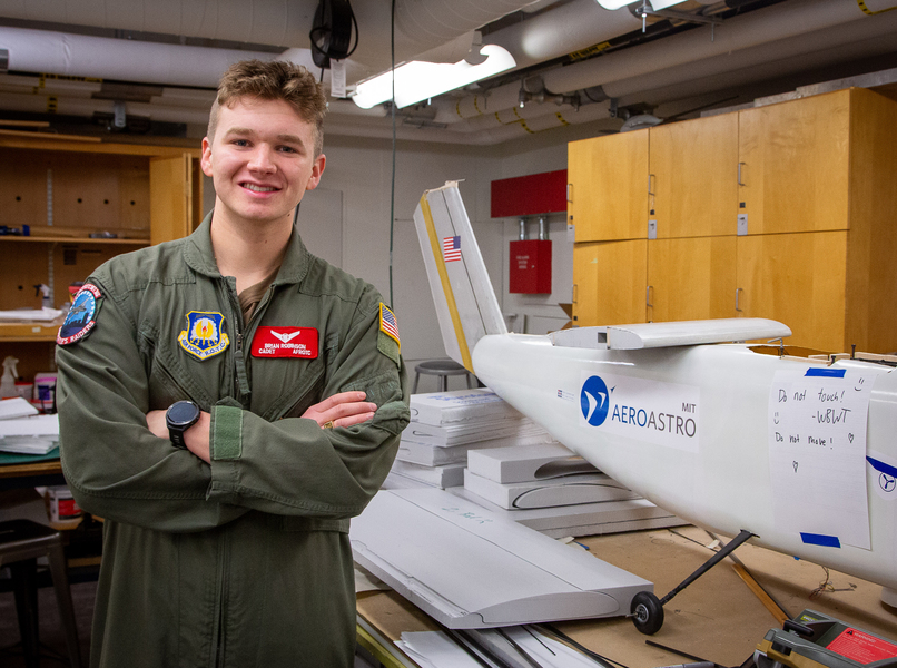 Brian Robinson, in an Air Force ROTC uniform, stands next to a model plane design with the MIT AeroAstro logo