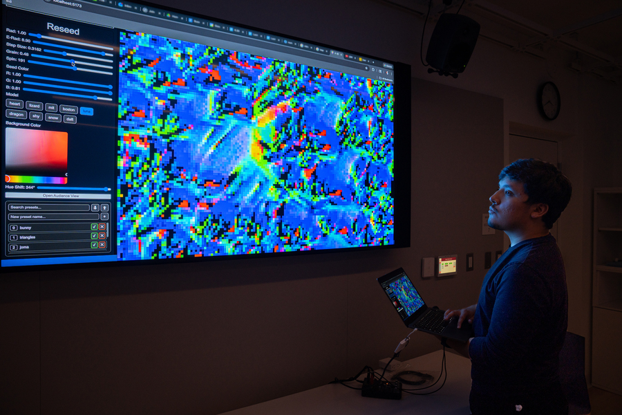 Carlos Mariano Salcedo stands in front of a large monitor displaying pixellated patterns in a darkened room. 