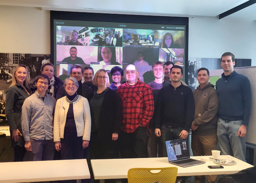 14 people stand around a large projection screen displaying at least a dozen other individuals on Zoom.