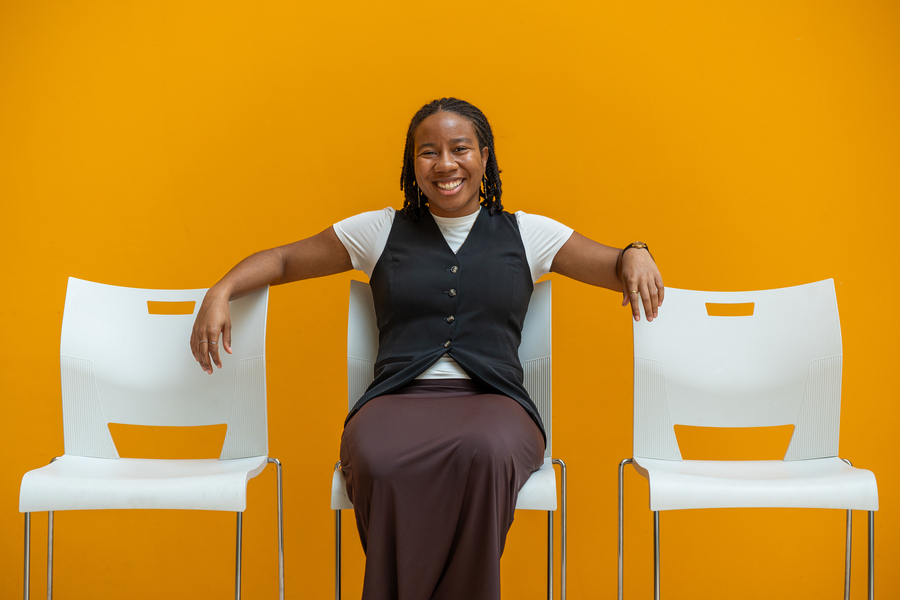 Olivia Honeycutt, a black woman with braids, poses seated on a white chair between two other white chairs before a golden yellow wall