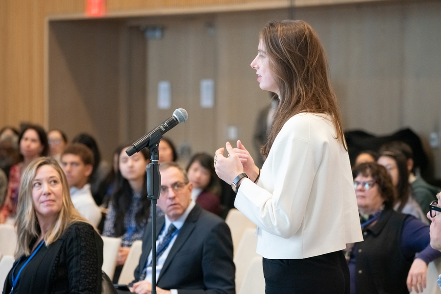 An audience member stands at a microphone. A full audience is visible seated behind her.