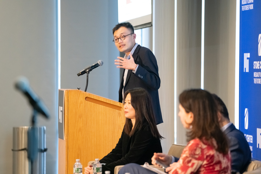 David Yang stands at a wooden MIT podium, gesturing while presenting at an indoor event.