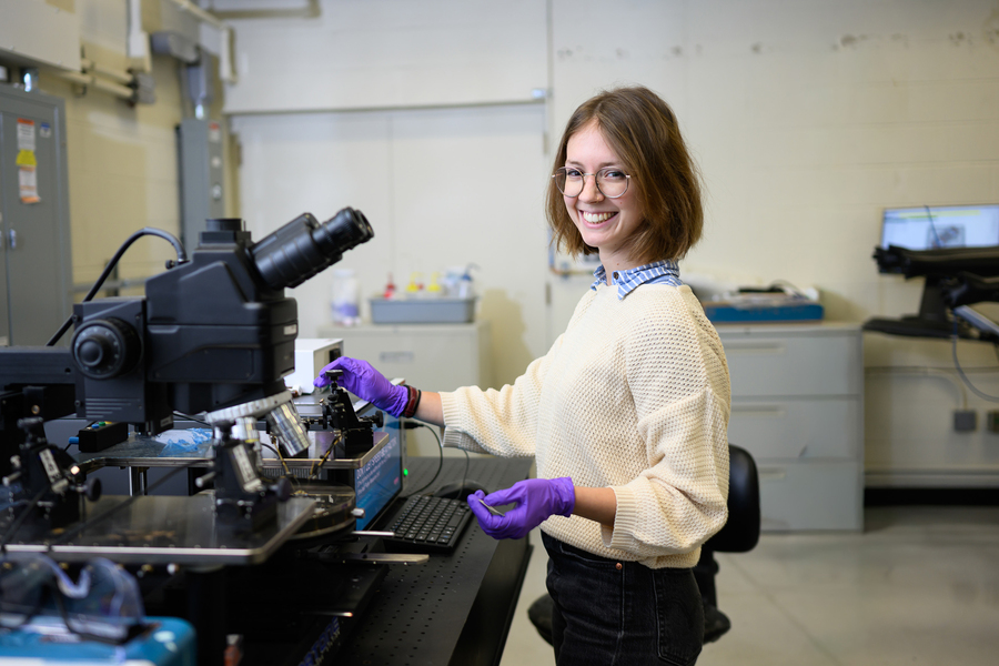 Camille Cunin stands at a laboratory worktable using a microscope. She wears purple nitrile gloves and smiles broadly at the camera.