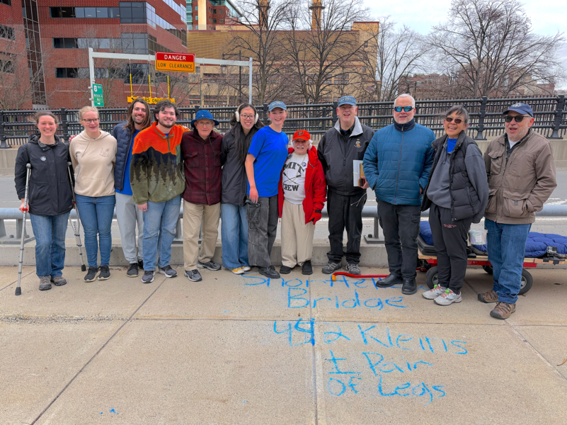12 people stand together at the end of a bridge with the writing, “Shortfellow Bridge - 442 Kleins + pair of legs” in chalk on the sidewalk.